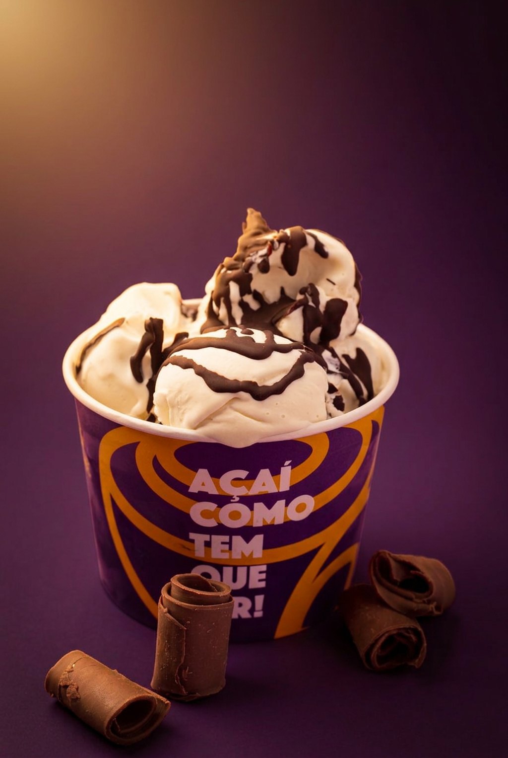 Pote AÇAIST com sorvete cremoso e calda de chocolate, acompanhado de rolinhos de chocolate, sobre fundo roxo profundo